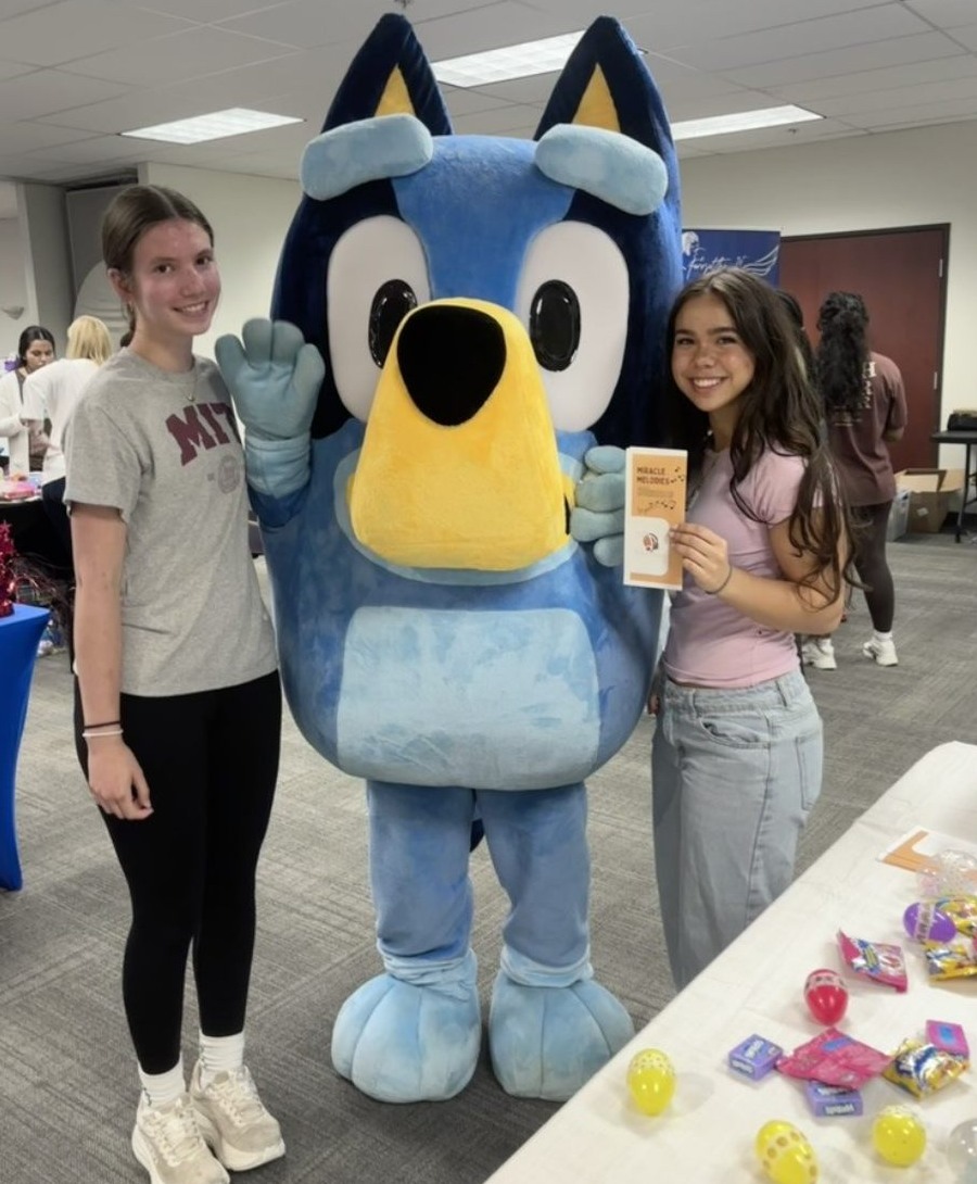 Two Miracle Melodies volunteers beside a costumed character at a family event, one holding a Miracle Melodies brochure.