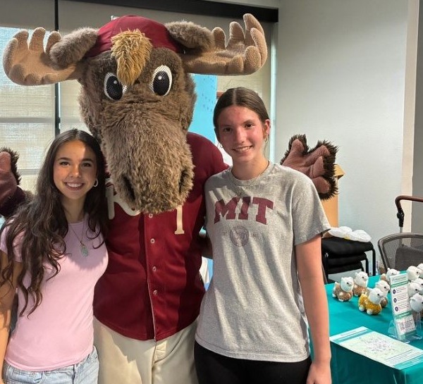 Two Miracle Melodies volunteers standing either side of a costumed moose mascot at a community event table.