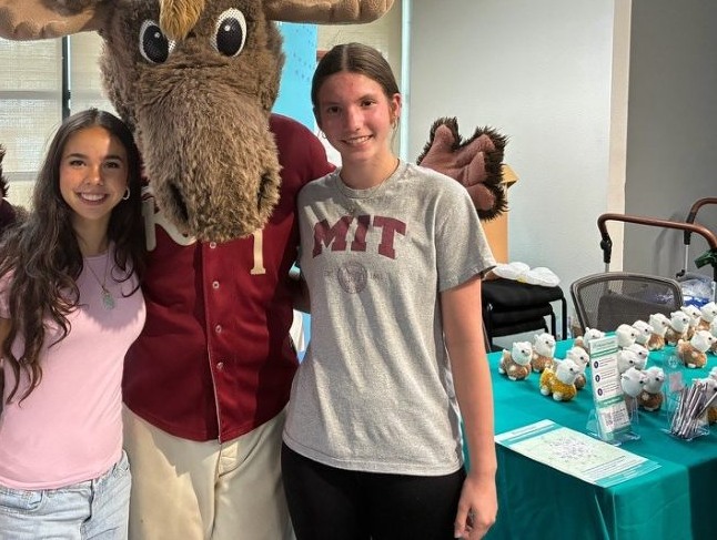 Two Miracle Melodies volunteers with a costumed moose mascot at a community outreach table.